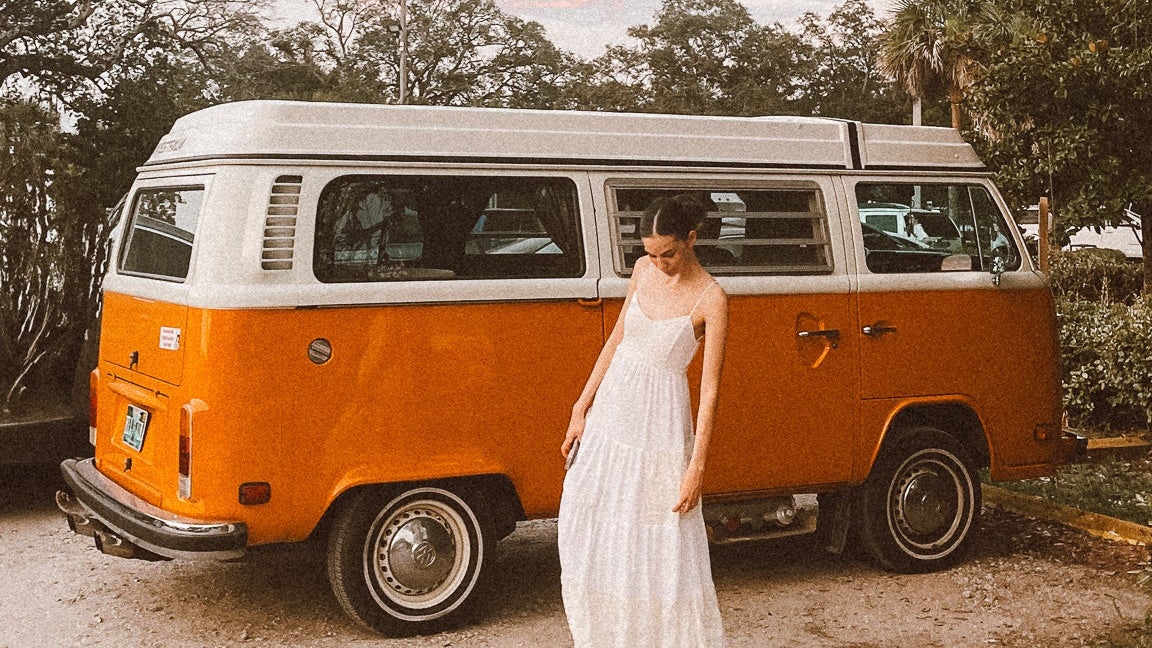 Woman in a white dress standing next to an orange and white van with trees and sky in the background